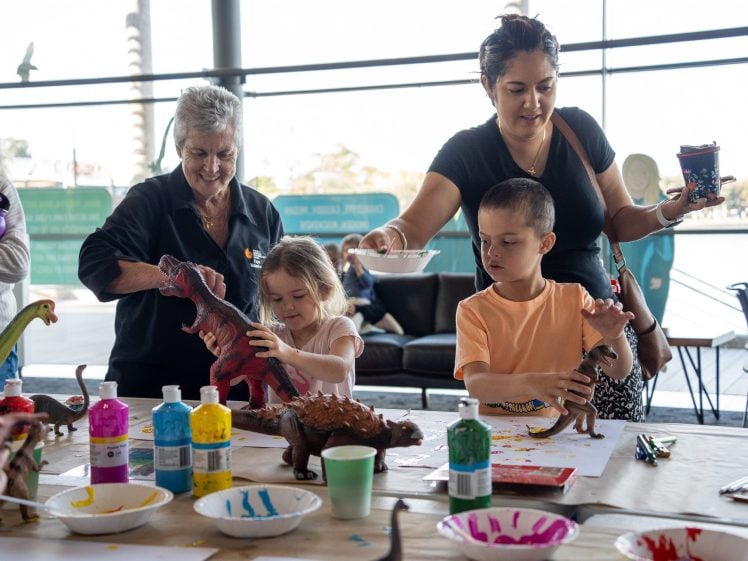 Image of An adult and two children paint dinosaur figurines at a table covered with art supplies, supervised by an older woman. Colourful paint bottles, brushes, and papers are spread out, and a window shows an outdoor view in the background.