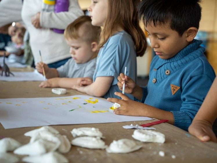 Image of Children sit at a table, painting and drawing on paper with brushes. Art supplies and clay pieces are scattered on the table. The children are focused on their creative activities in a bright indoor setting.