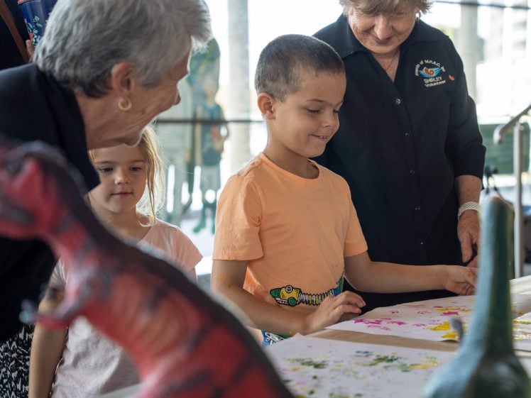 Image of A young boy and girl are doing an art activity at a table, watched by two older women. Colourful dinosaur toys and paint are visible on the table. Everyone looks engaged and happy.