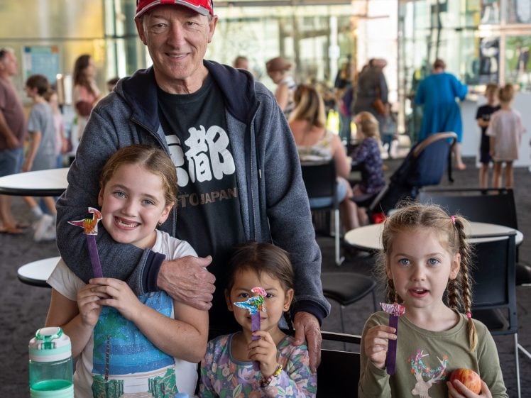 Image of An older man stands behind three smiling young girls at a crowded indoor venue. The girls hold colourful drink stirrers and snacks. People are seated and walking in the background.
