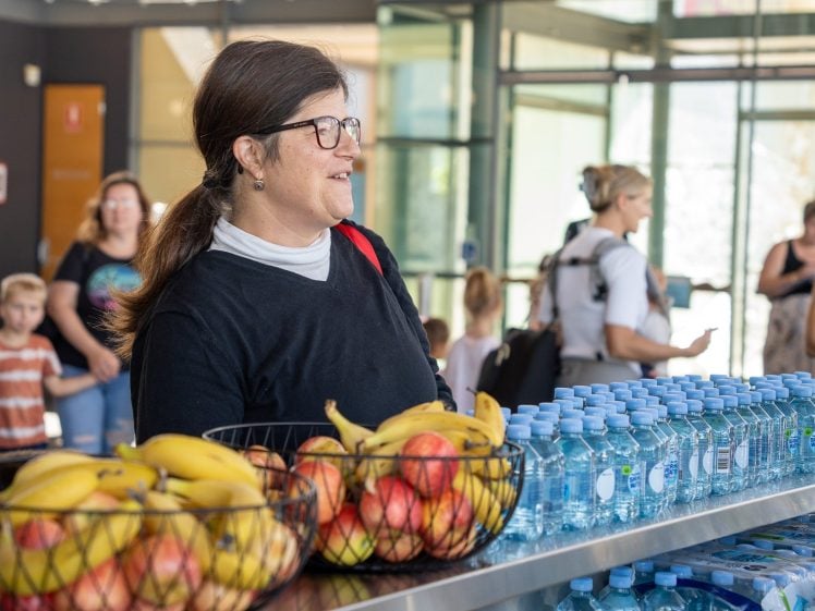 Image of A woman with glasses stands by a table with baskets of bananas and apples, and rows of bottled water. Other people, including children, are visible in the background of the bright indoor setting.