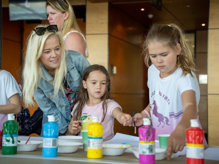 Image of A woman and two young girls are at a table with colourful paint bottles and bowls. One girl paints while the other, guided by the woman, looks at her artwork. People are in the background in a bright indoor setting.