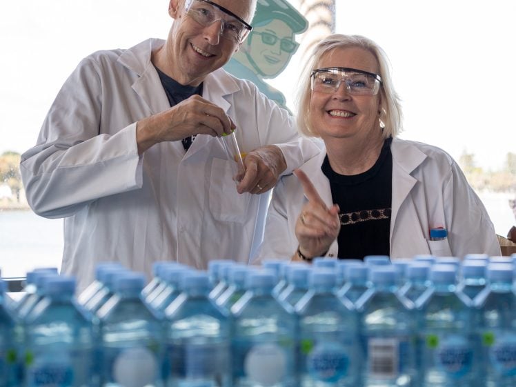 Image of Two smiling scientists in lab coats and safety glasses stand behind rows of water bottles. One holds a pipette and the other points forward. There is a waterfront scene visible in the background.