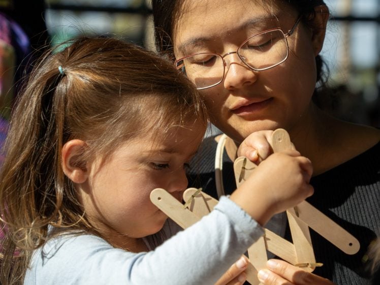 Image of A woman with glasses helps a young girl build a structure with wooden craft sticks. Sunlight shines on their faces as they focus closely on the creative activity together.