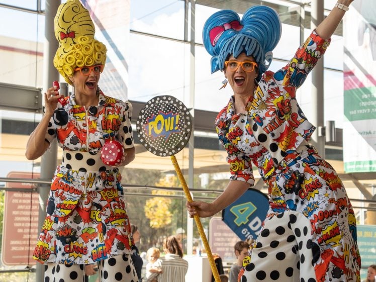 Image of Two women dressed in colourful comic-themed outfits and large whimsical wigs pose playfully indoors. One holds a polka-dot POW! sign; both wear bright glasses and polka dot trousers, entertaining people in the background.