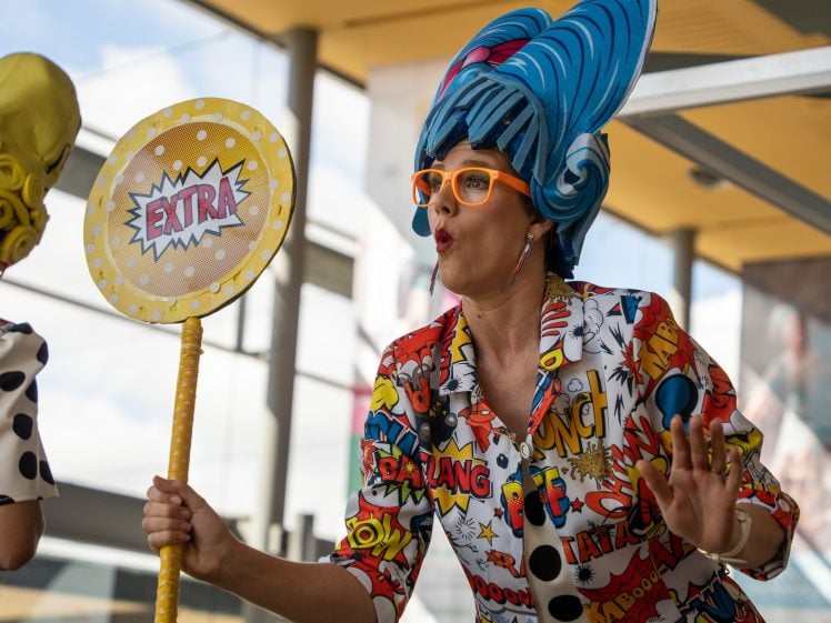 Image of A person dressed in a colourful comic-themed outfit and blue wig holds a yellow sign that says EXTRA. They wear orange glasses and pose expressively indoors with modern glass architecture in the background.