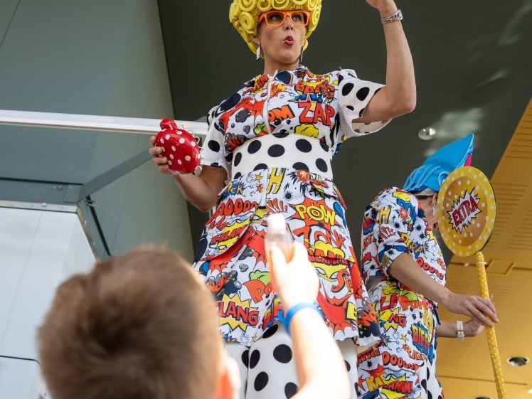 Image of A performer in a colourful, comic-themed costume and yellow wig stands on stilts, blowing bubbles for a child reaching up in a bright indoor setting.