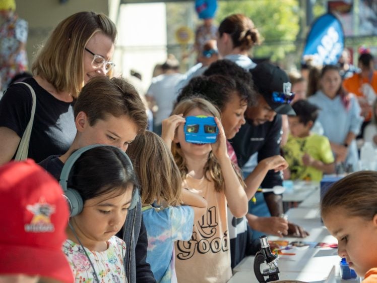 Image of Children and adults gather around a table at a busy indoor event, exploring science activities. A girl uses a blue viewer, another looks through a microscope, and others engage with interactive displays. The scene is lively and bright.