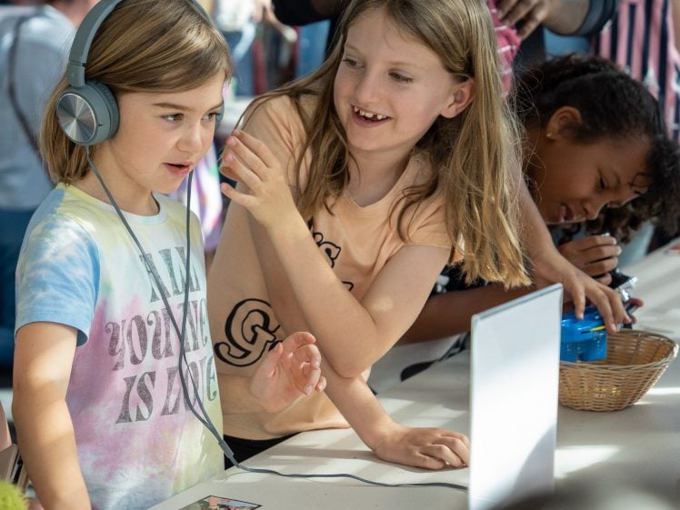 Image of Two young girls interact at a table with photos and a laptop; one wears headphones and a tie-dye shirt, whilst the other smiles and gestures. Another child is in the background with a blue toy.
