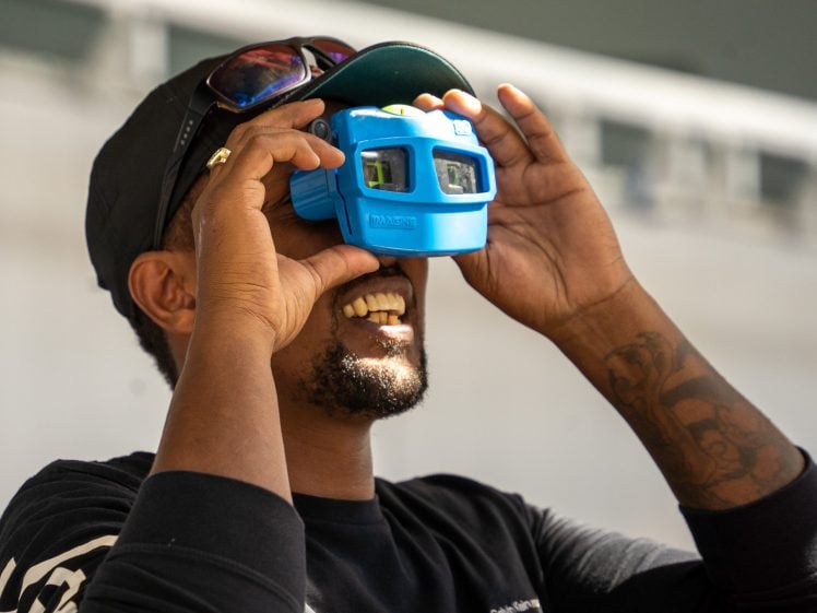 Image of A smiling man wearing a cap and sunglasses on his head holds a blue View-Master toy viewer to his eyes, looking up. He has a visible tattoo on his forearm and a trimmed beard.