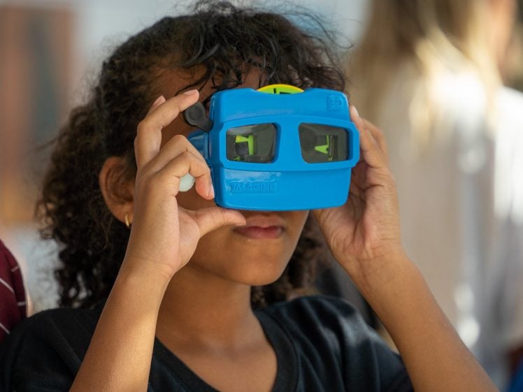 Image of A child with curly hair holds a blue stereoscope viewer up to their eyes, looking through it with curiosity. The background is softly blurred.