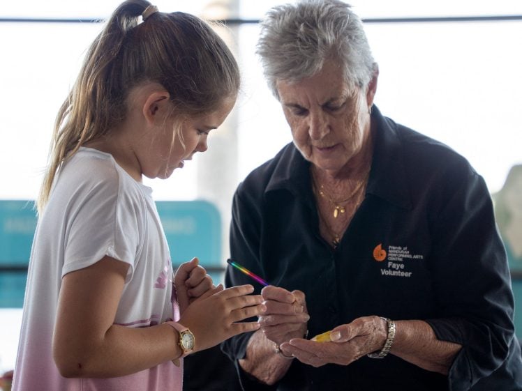 Image of An older woman wearing a “volunteer” shirt paints the palm of a young girl’s hand while the girl watches, indoors with bright natural light in the background.