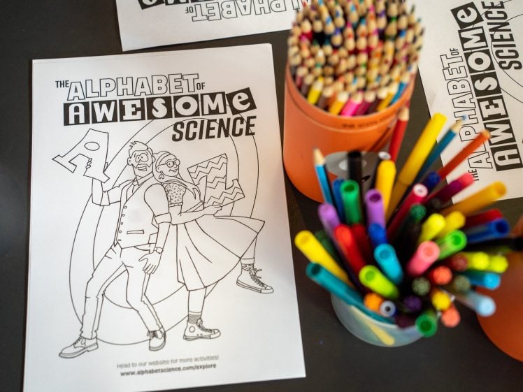 Image of A colouring page titled The Alphabet of Awesome Science lies on a table with illustrated scientists, surrounded by coloured pencils, markers, and a cup of coffee.