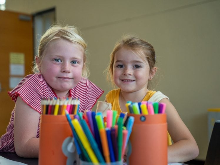 Image of Two young girls smile at a table covered with colourful pencils and markers in containers, with a blurred classroom background.