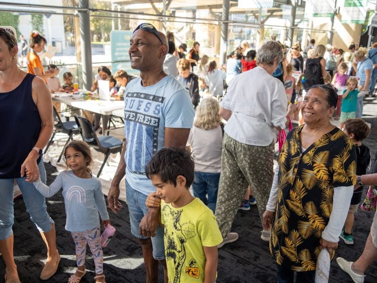 Image of A diverse group of adults and children gather indoors at a community event, smiling and interacting in a bright, open space with tables and large windows in the background.