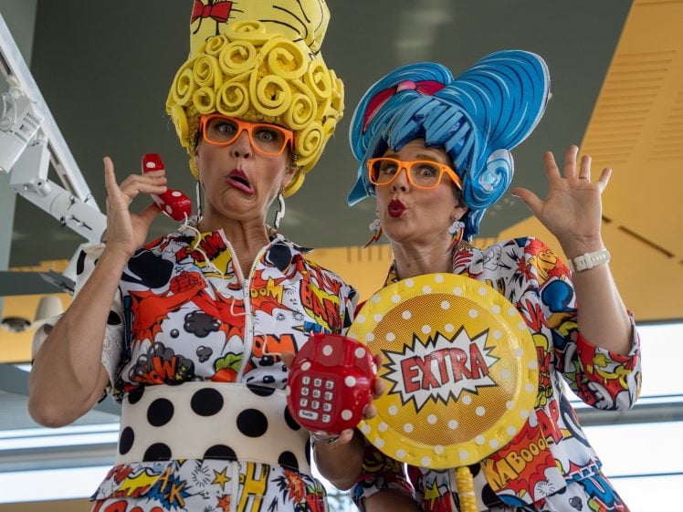 Image of Two people dressed in colourful, comic-themed outfits and oversized, cartoonish wigs pose playfully. One holds a red toy phone; the other holds a large, round prop with EXTRA on it. Both wear orange glasses and pull funny faces.