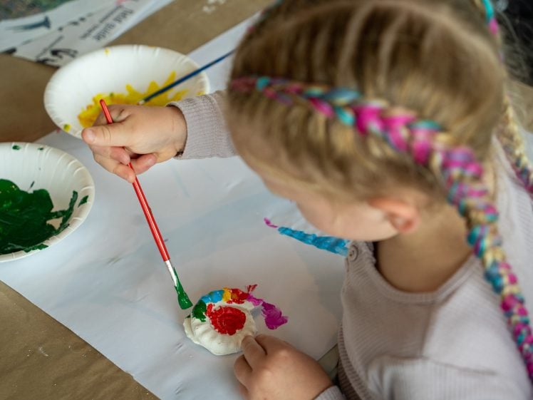Image of A young child with colourful braided hair uses a red paintbrush to paint a clay ornament, with bowls of green and yellow paint nearby on a table covered with paper.