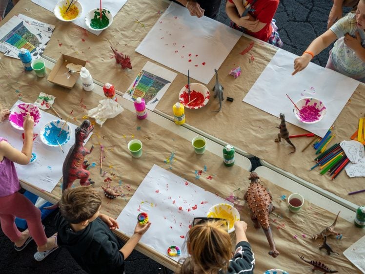 Image of Children gather round a table covered with paper, paint, brushes, toy dinosaurs, and art supplies, creating colourful artwork together in a lively and creative setting.