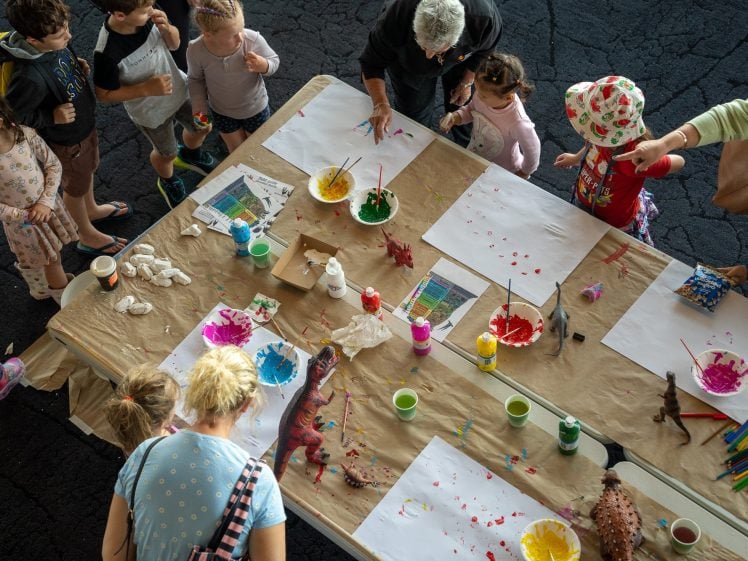 Image of Children and adults gather round a table covered with brown paper, art supplies, paint, and dinosaur toys, creating colourful paintings at a group art activity. Some children are painting, while others watch.