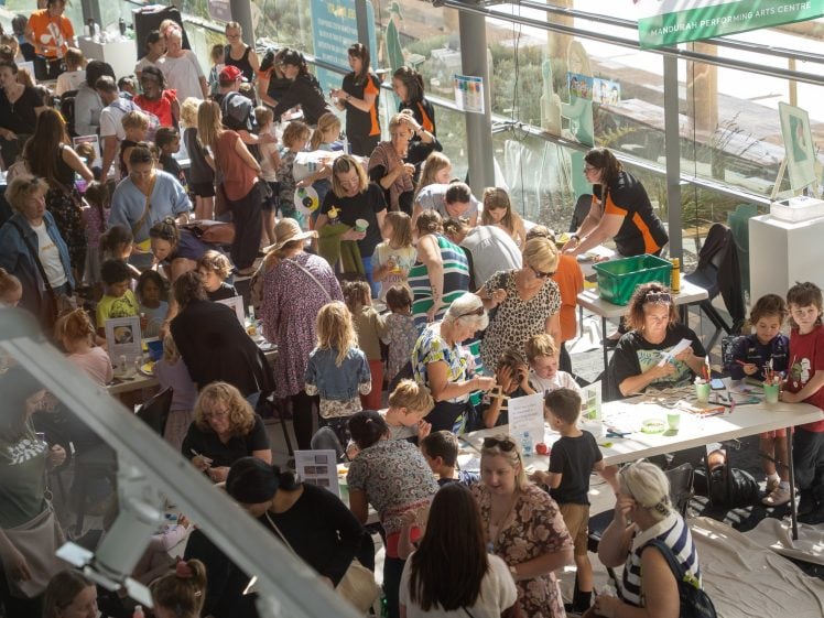 Image of A busy indoor event with many adults and children gathered around tables, engaging in arts and crafts activities under bright natural light from large windows. Various materials and art supplies are spread out on the tables.