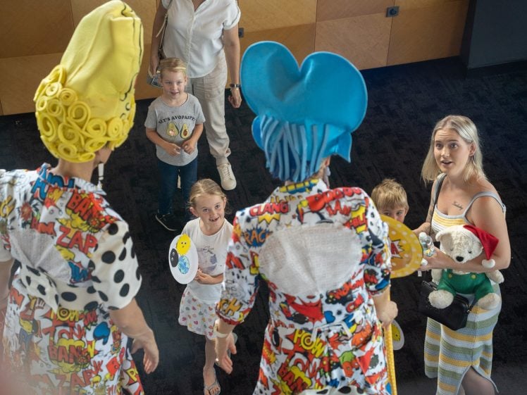 Image of Two costumed performers with bright yellow and blue wigs face a smiling group of adults and children indoors. The performers wear colourful, comic book–themed outfits. The children hold fans with smiley faces.