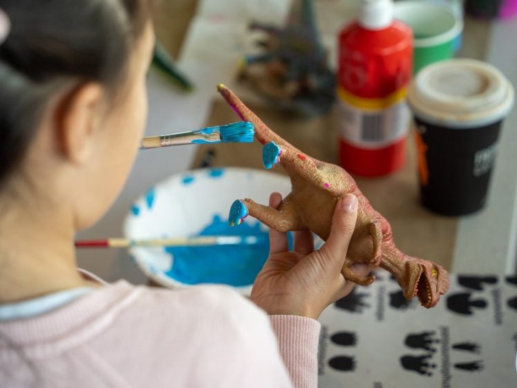 Image of A child uses a paintbrush to paint blue spots on a toy dinosaur. Nearby are bottles of paint, a bowl of blue paint, and a cup, on a table covered with art supplies.