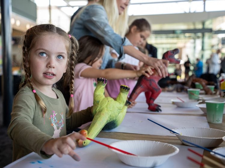Image of A young girl with plaited hair holds a green dinosaur toy and a red paintbrush at a table with other children, painting dinosaur models. Adults assist in the background in a bright indoor space.