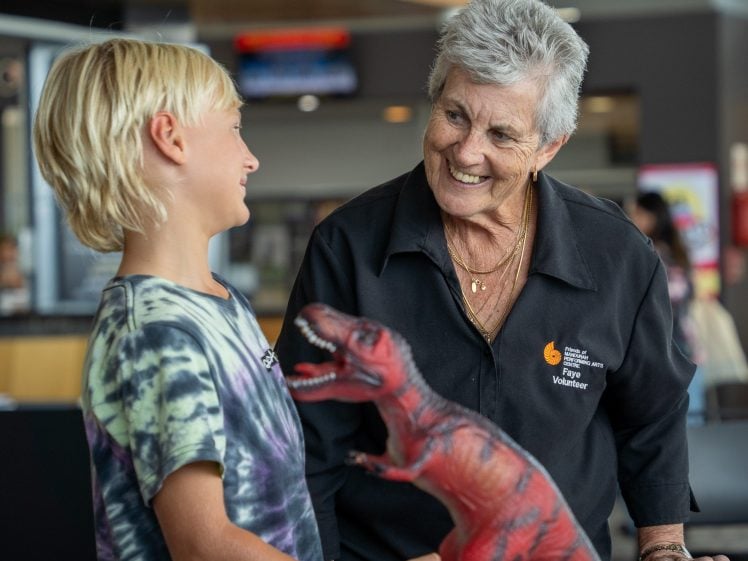 Image of An elderly woman and a young boy smile at each other whilst standing at a table with a red toy dinosaur. The woman wears a volunteer shirt, and they appear to be at an indoor community or educational event.