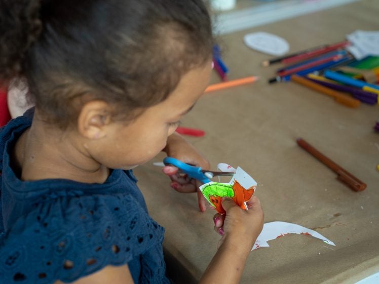 Image of A young child uses blue scissors to cut out a colourful drawing on white paper at a table scattered with coloured pencils and art supplies.