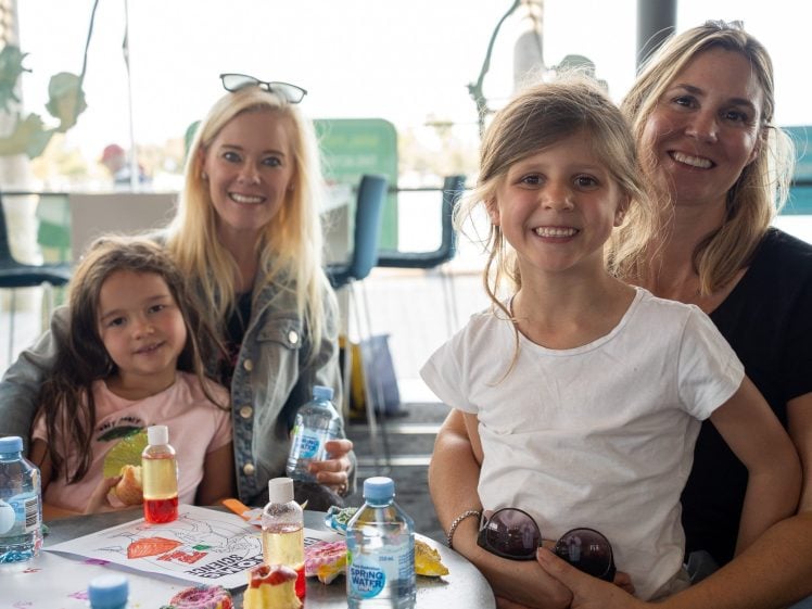 Image of Four people, two adults and two children, sit at a table with drinks, snacks, and colouring pages. All are smiling at the camera in a bright indoor setting with large windows in the background.