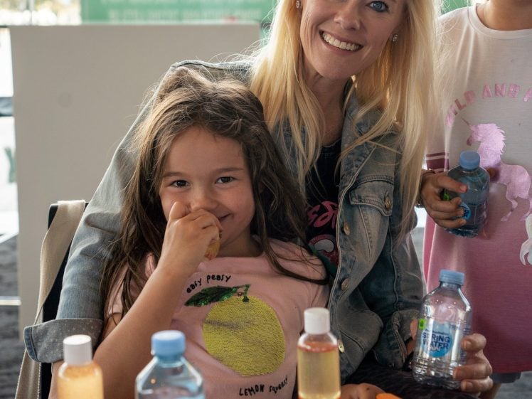 Image of A smiling woman sits with two young girls at a table covered with drawings, felt-tip pens, and bottles of coloured liquid. One girl eats a snack, while the other holds a blue drink. They appear to be enjoying an arts and crafts activity.