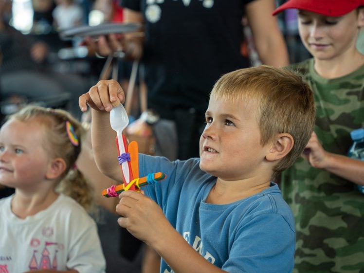 Image of A young boy holds up a homemade catapult made from lolly sticks and a plastic spoon, surrounded by other smiling children at an indoor event.
