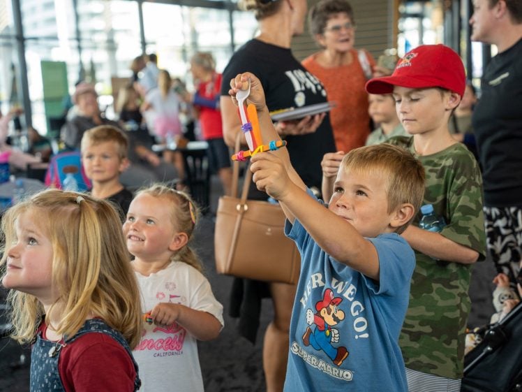 Image of A group of young children and adults gather indoors. A smiling boy at the front, wearing a blue shirt, holds up a colourful craft project. Other children watch with interest and joy, while adults look on in the background.