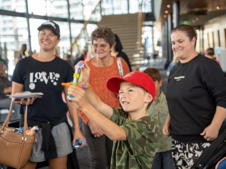 Image of A young boy in a red cap plays with a colourful toy, smiling as three women and a child behind him watch and laugh in a spacious indoor setting with large windows and stairs in the background.