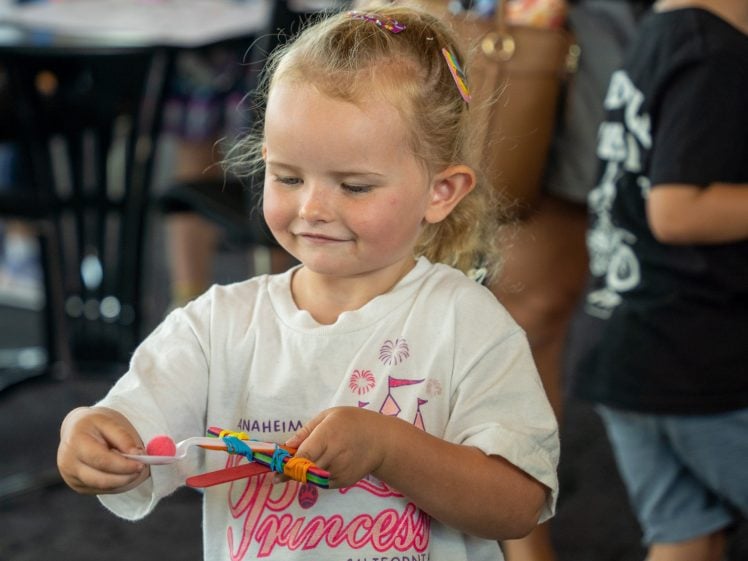Image of A young girl with blonde hair holds a lolly-stick craft with a pink pom-pom. She is smiling and wearing a white Princess California T-shirt. Other people are blurred in the background.