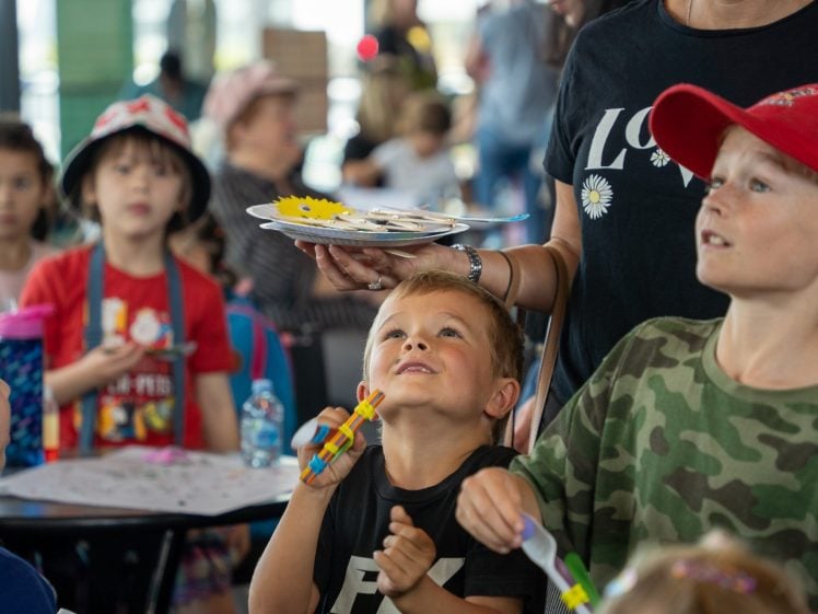 Image of A group of children at a table doing crafts, with one child looking up and smiling at a plate of materials held by an adult. The setting appears busy and cheerful with other children in the background.