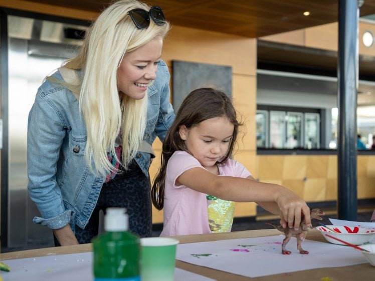 Image of A smiling woman with long blonde hair watches a young girl press a toy dinosaur onto paper, creating colourful prints in a bright indoor space, with art supplies on the table.