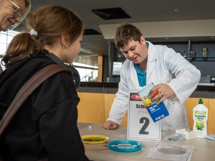 Image of A scientist in a white lab coat leads a science activity at a table, showing a carton to a young girl. Plates and washing-up liquid are on the table, and a Science sign with the number 2 is displayed.