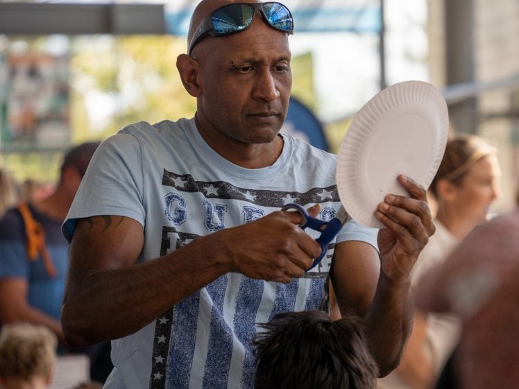 Image of A man in a light blue shirt concentrates as he cuts a white paper plate with scissors at a busy indoor event. People and children are visible in the background.