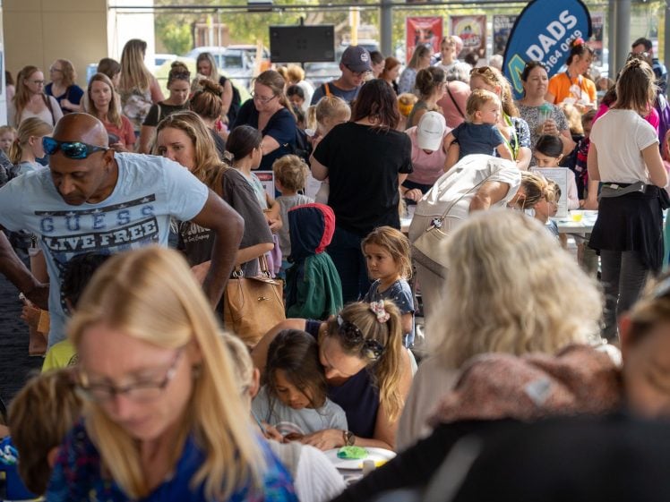 Image of A busy indoor event with many adults and children gathered around tables, engaging in activities and crafts. The room is crowded and lively, with people talking and working together.