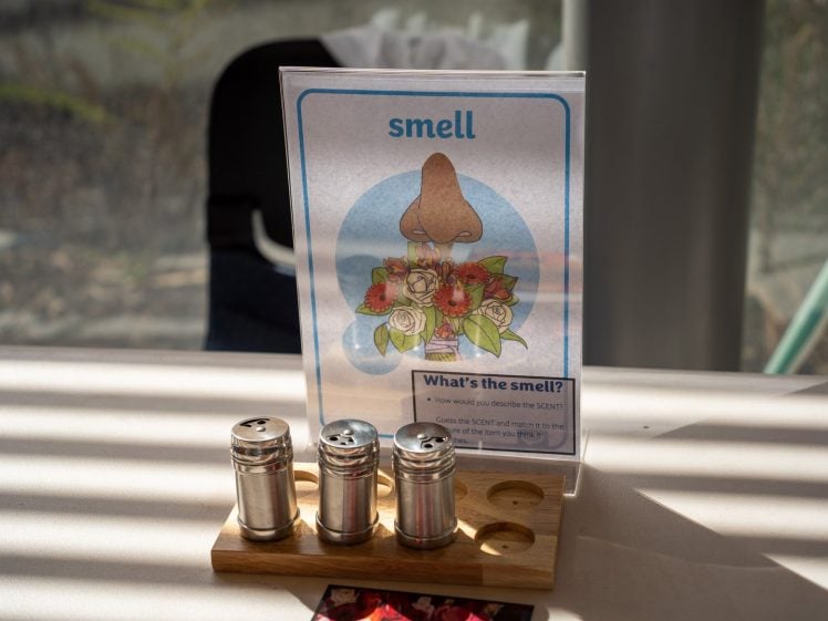 Image of A sign with an illustration of a nose smelling flowers is labelled “smell.” In front of the sign are three metal shakers on a wooden holder, used as part of a scent activity. Sunlight and shadows fall across the table.