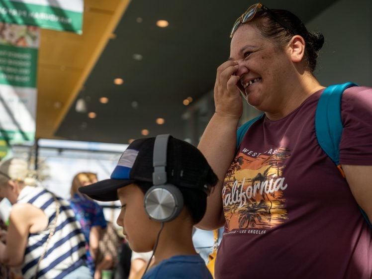 Image of A smiling woman wearing a California T-shirt and blue rucksack stands next to a boy in headphones and a cap. They are indoors near other people, with sunlight streaming in and green signs hanging above.