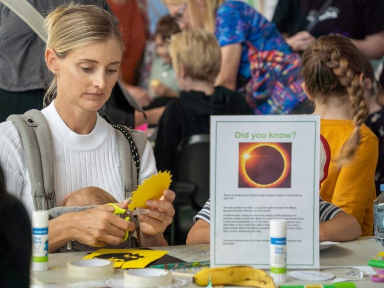 Image of A woman holds a baby while cutting yellow paper at a craft table. A sign with an eclipse image and “Did you know?” text is on the table, along with glue sticks, craft supplies, and a banana. Other people are crafting in the background.