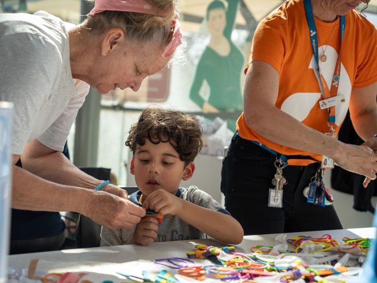 Image of An older woman helps a young boy with a colourful craft project at a table, while another adult wearing an orange shirt and lanyard organises supplies nearby. The table is covered with colourful craft materials.