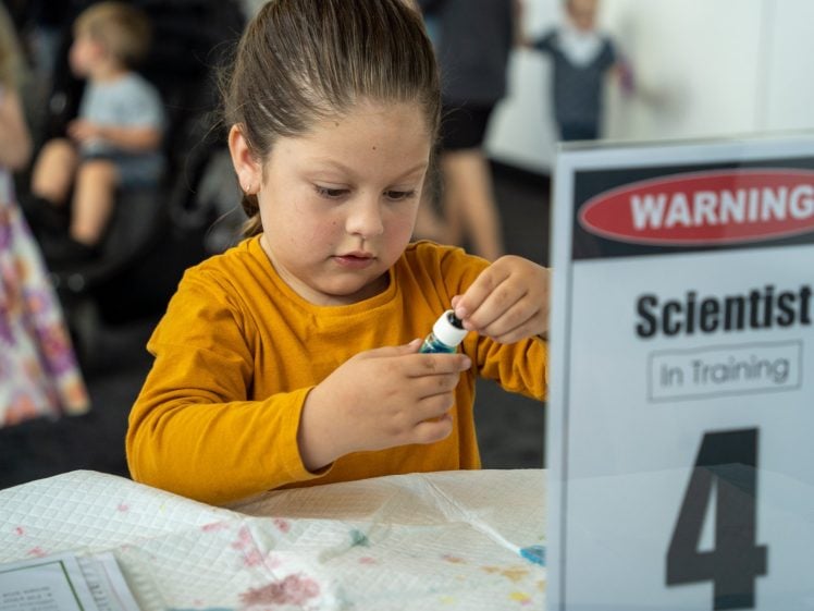 Image of A young child in a yellow shirt concentrates whilst holding a small vial at a table covered with a white cloth. A sign nearby reads “Warning! Scientist In Training 4.” Other children are visible in the background.