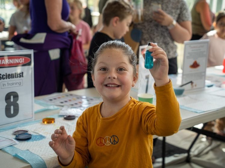 Image of A young child in a yellow shirt smiles and holds up a small blue phial at a science activity table. Other children and adults are engaged in experiments in the background. A sign reads “Scientist in Training”.