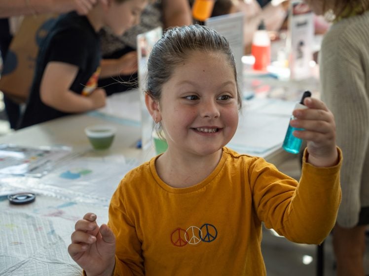 Image of A young girl in a yellow shirt with peace signs smiles and holds up a small bottle of blue liquid at a busy table filled with papers and craft supplies. Other children work in the background.
