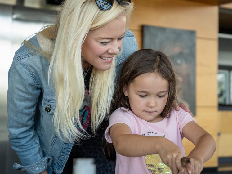 Image of A woman with blonde hair smiles and watches as a young girl uses her fingers to paint on paper at a table, with art supplies nearby in a bright indoor setting.