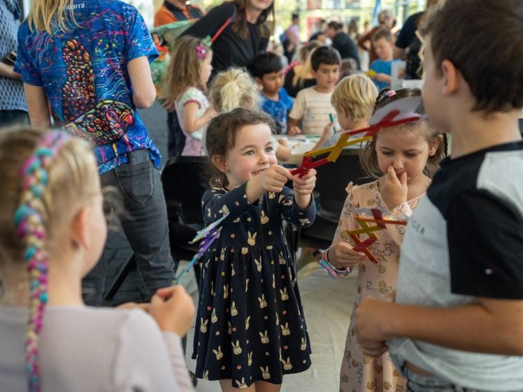 Image of A group of young children are gathered indoors, smiling and playing with colourful craft sticks. One girl in a dark dress with bunny prints holds up her craft towards a boy, while other kids look on and join the fun.