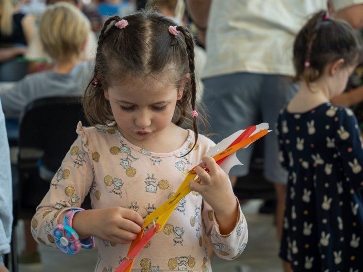 Image of A young girl with plaits and a floral dress concentrates on crafting with coloured paper in a busy, indoor setting with other children nearby.